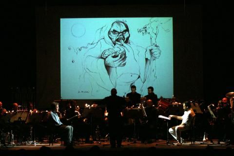 Fotografia di scena: un'orchestra suona sul palco di un teatro, il direttore di spalle al centro, e sopra di loro un grande schermo proietta il disegno a china di una figura mostruosa.